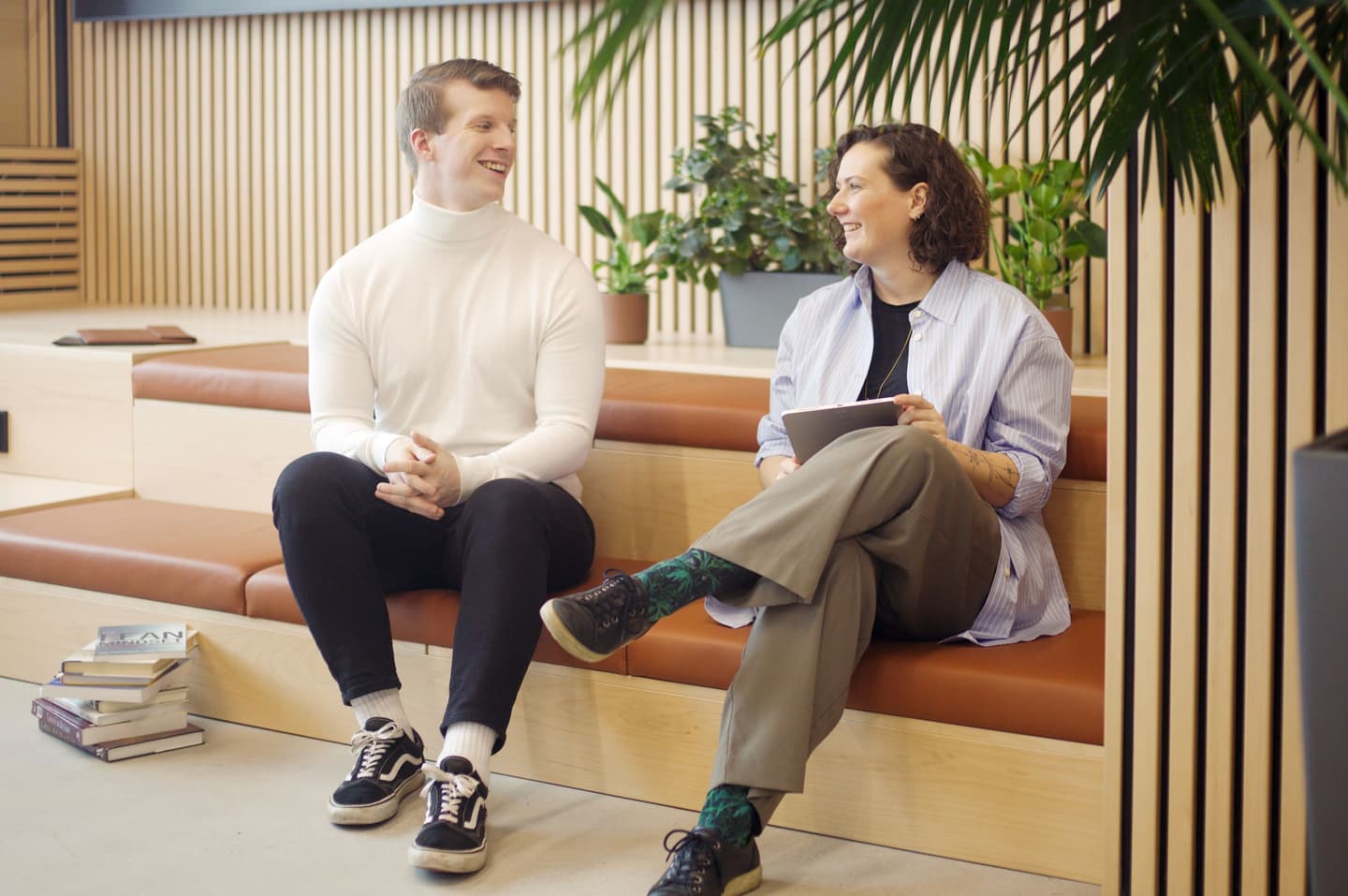 Two people having a friendly conversation on wooden seating steps in a modern office space with plants in the background.