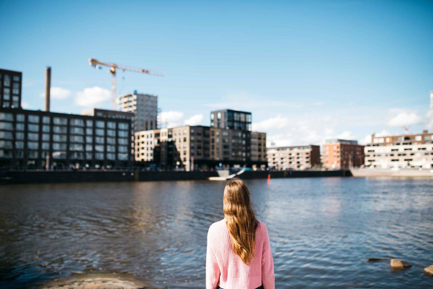 Person in pink jumper looking across urban waterfront with modern apartment buildings and construction crane under blue sky.