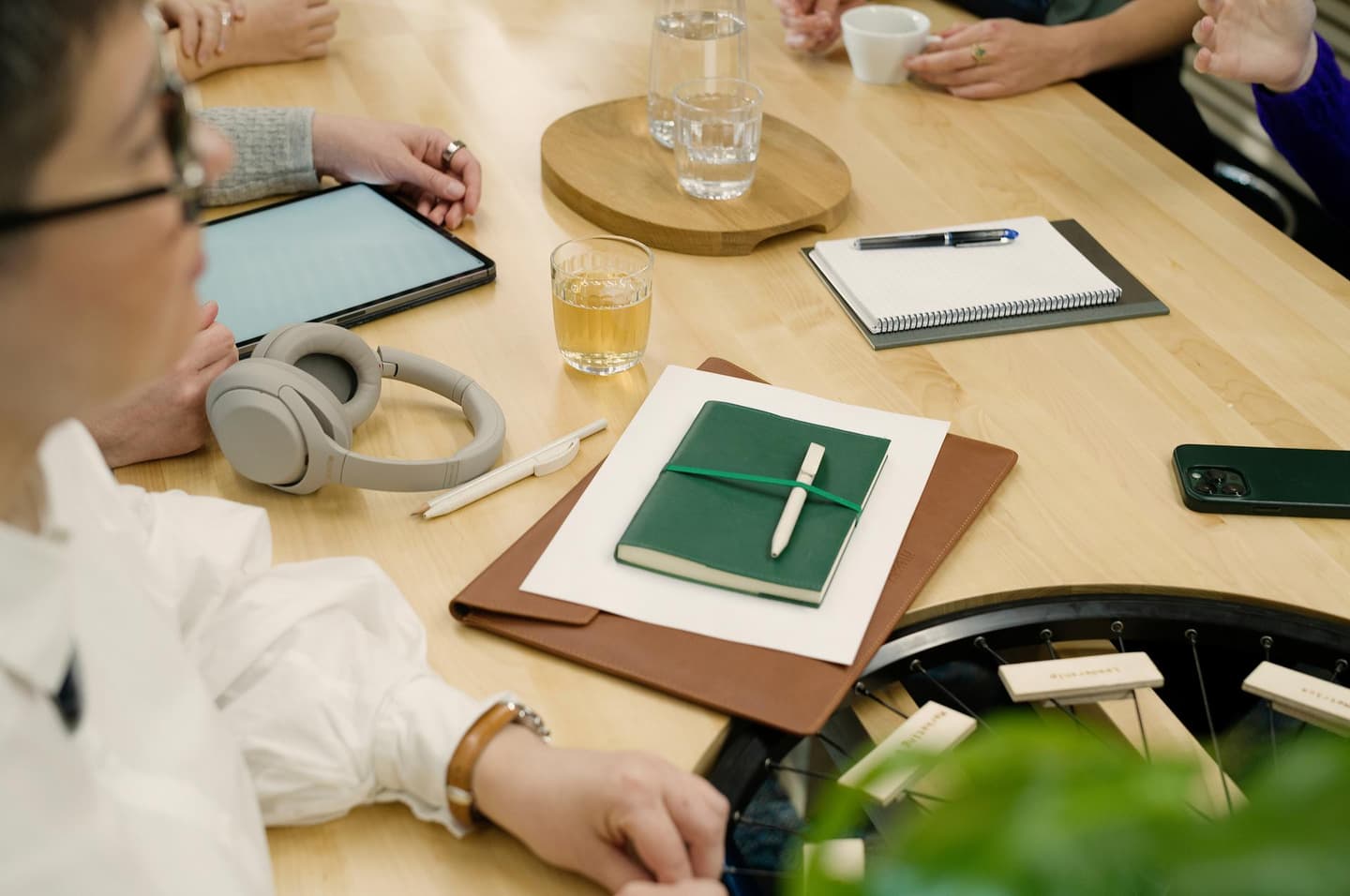 People gathered around wooden table with tablet, headphones, notebooks, drinks and stationery during a meeting.