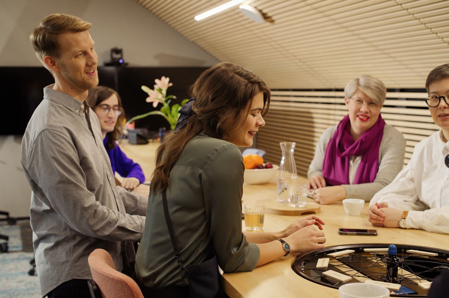 A diverse group gathered around a wooden table with drinks, engaged in conversation in a modern space with wooden ceiling.