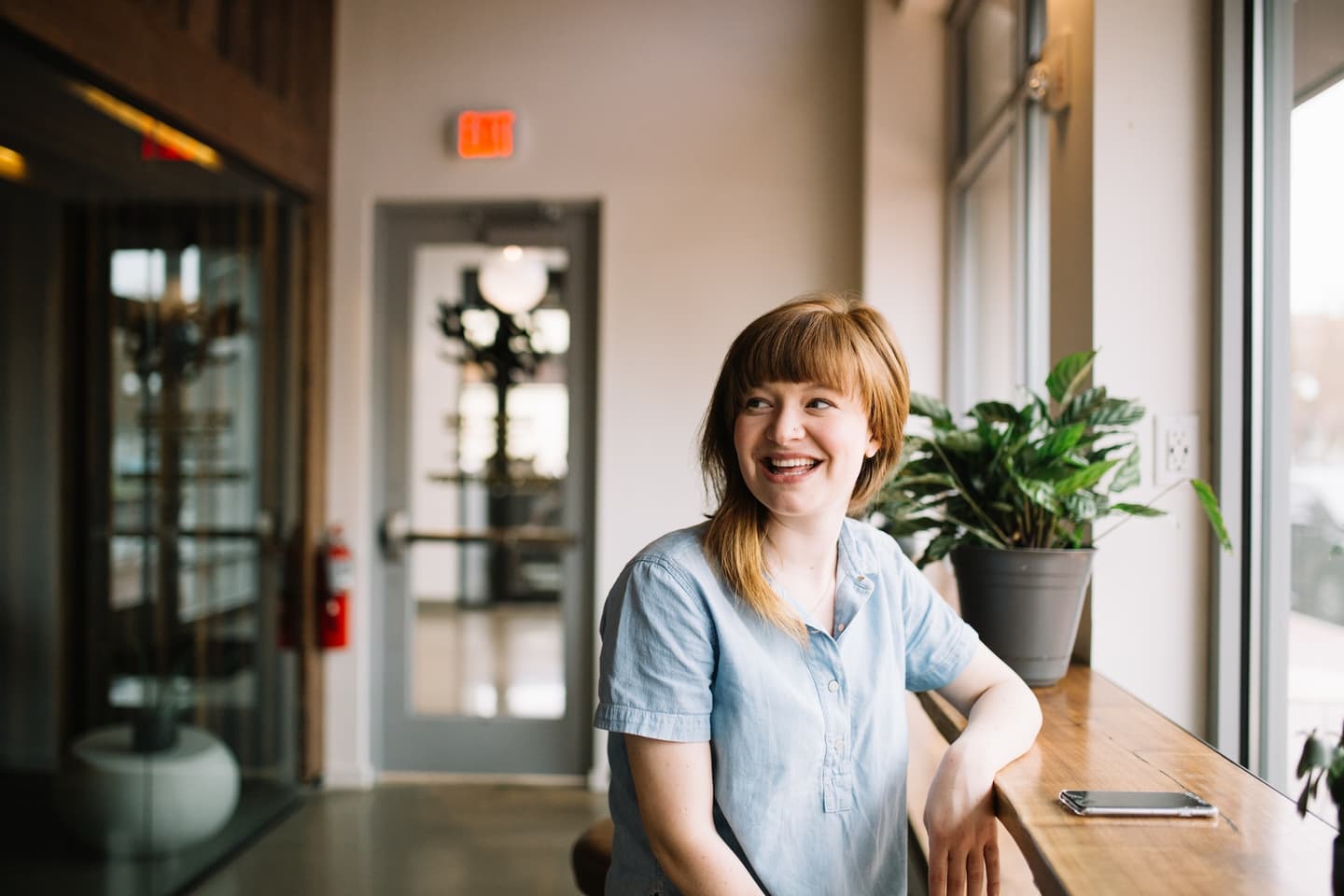 Person with auburn hair in light blue shirt smiling by window with potted plant in bright modern office space.