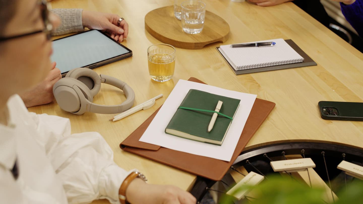 Wooden table with meeting items: tablet, headphones, green notebook, notepad, drinks, and people collaborating.