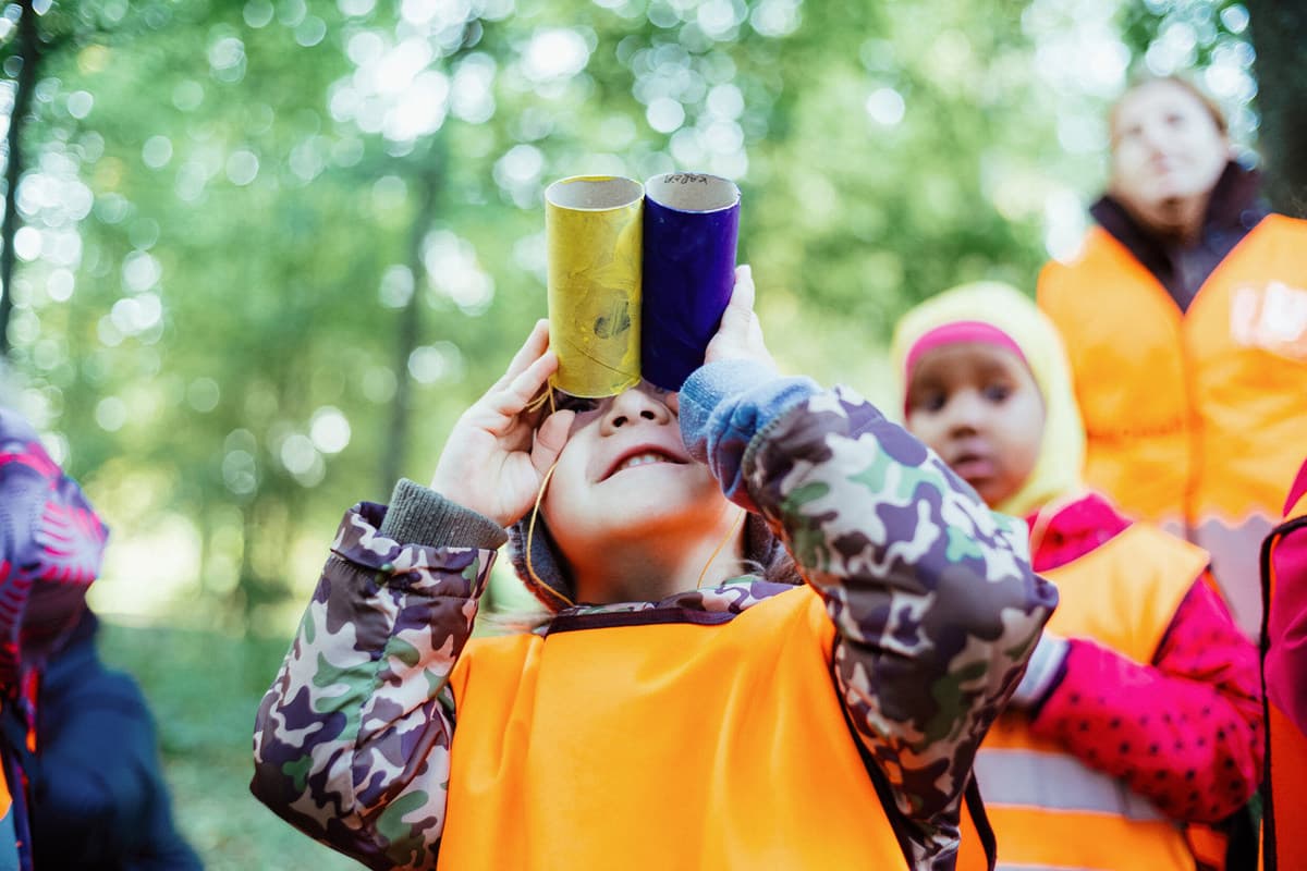 Child in orange safety vest looking through cardboard binoculars during outdoor forest activity with classmates.