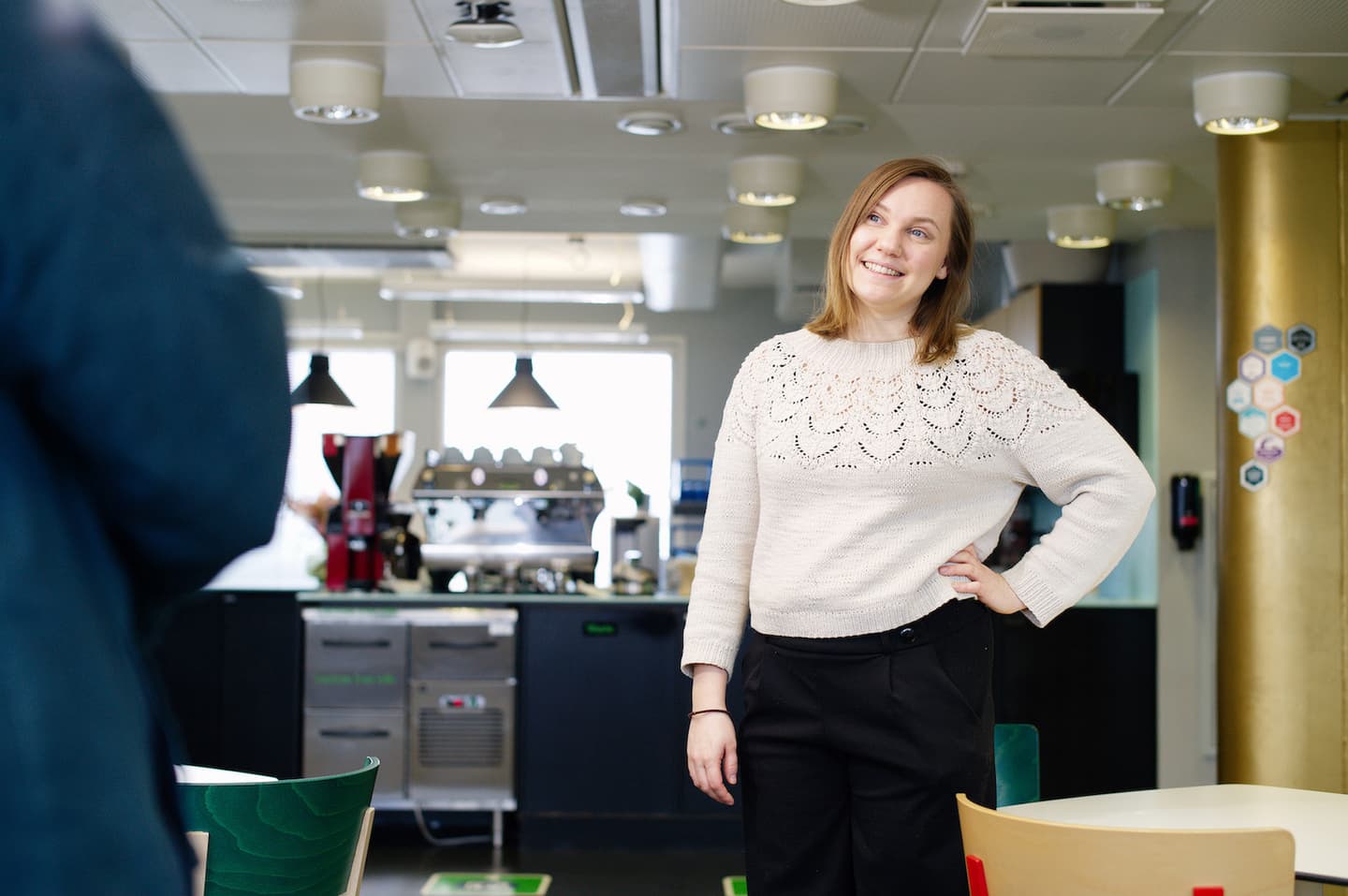 Person in white knitted jumper smiling in modern office break room with coffee area in background.